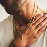 Man wearing a gold chain with an angel wing pendant, holding his hand to his chest.