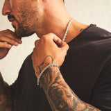 Man with tattoos wearing a silver chain necklace and bracelet against a neutral background